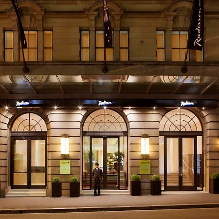Radisson Blu Plaza Hotel Sydney Eksteriør billede The photo depicts the entrance of a Radisson hotel. The building features elegant architecture, with large arched windows and a well-lit canopy above the entrance. There are two potted plants on either side of the doors, which are framed by modern li