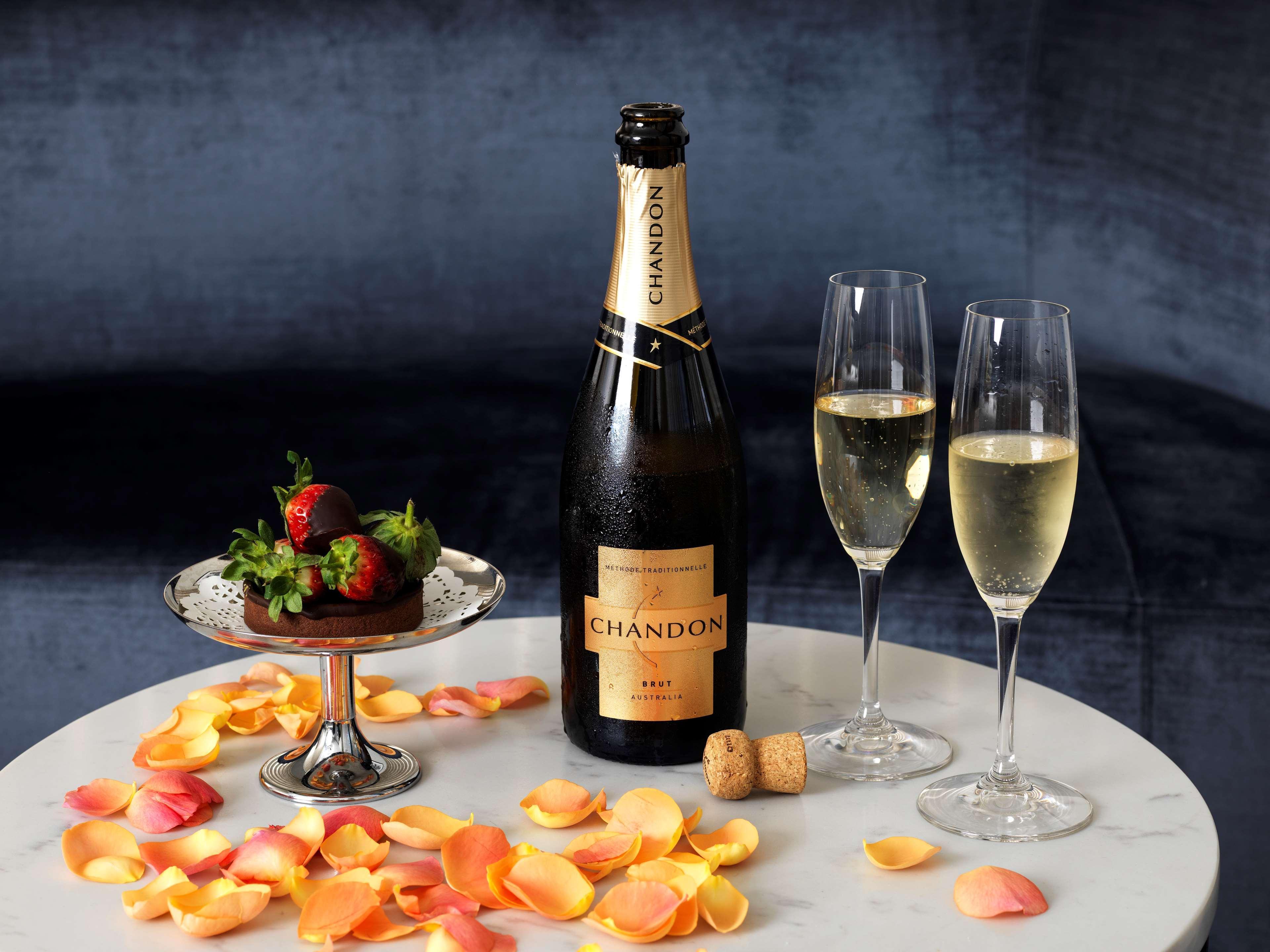 Radisson Blu Plaza Hotel Sydney Eksteriør billede The image shows a stylish setting featuring a bottle of Chandon sparkling wine accompanied by two filled champagne flutes. There is a silver plate with a dessert topped with strawberries and some scattered flower petals on the table, creating a roman