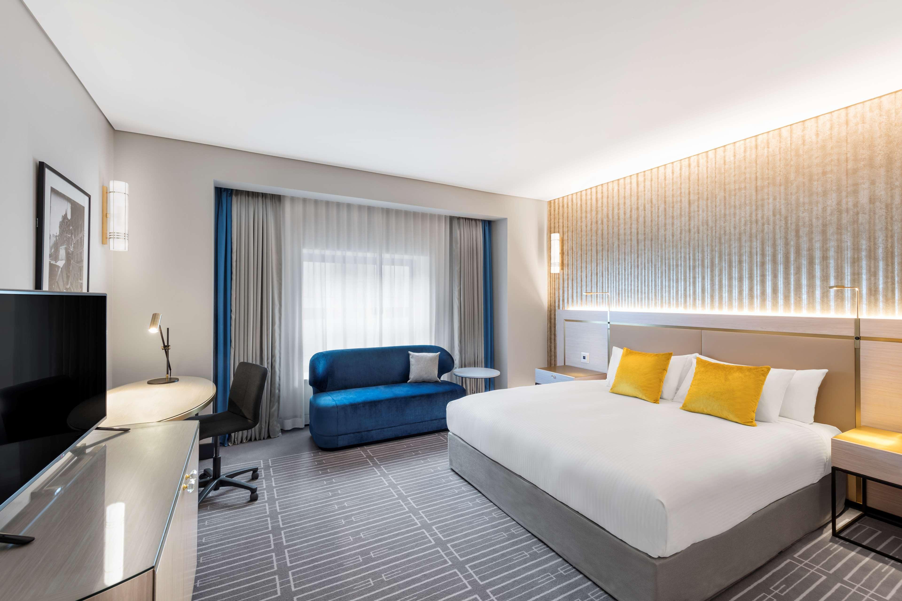 Radisson Blu Plaza Hotel Sydney Eksteriør billede The photo shows a modern hotel room featuring a spacious layout. It has a large bed with white linens and yellow accent pillows. Beside the bed, there is a bedside table and a contemporary lamp. There is a blue sofa and a coffee table near a set of l