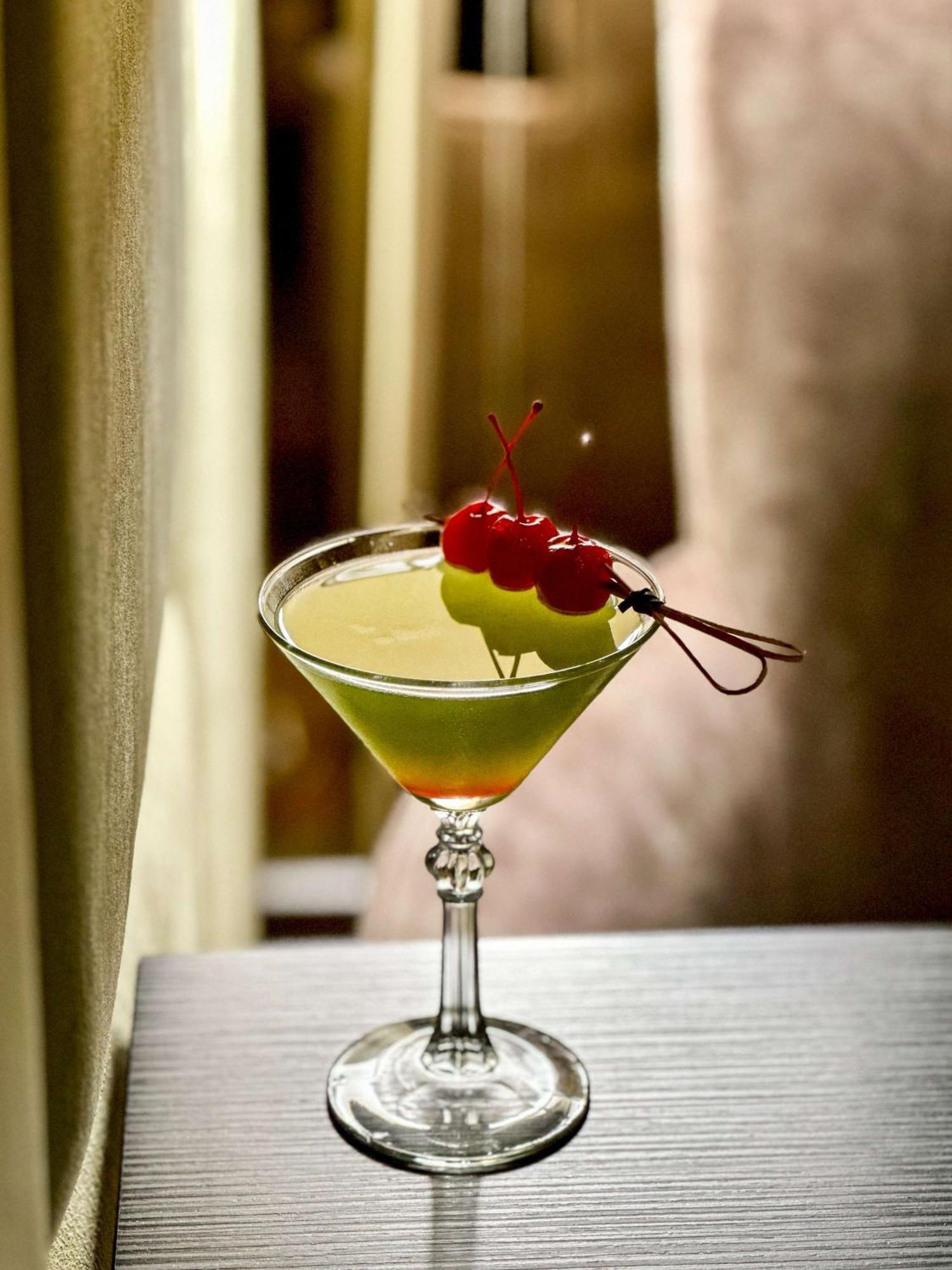 Radisson Blu Plaza Hotel Sydney Eksteriør billede The image shows a cocktail in a classic martini glass. The drink has a vibrant green color and is garnished with two yellow-green fruits (possibly a melon or similar) and three cherries on a skewer. The background appears blurred, creating a soft foc