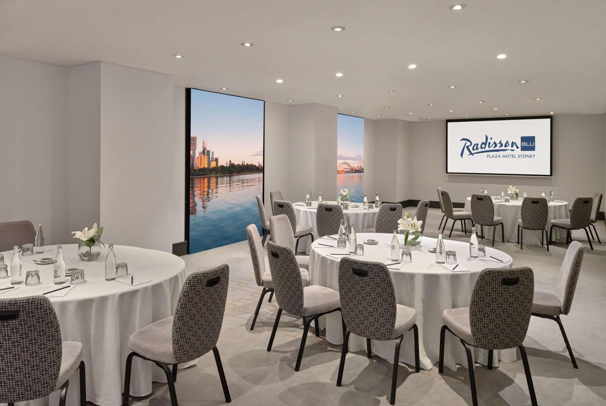 Radisson Blu Plaza Hotel Sydney Eksteriør billede The photo shows a modern meeting room at the Radisson Hotel in a city. The room features several round tables set with white tablecloths and gray upholstered chairs. There are small flower arrangements on each table, creating a welcoming atmosphere. 